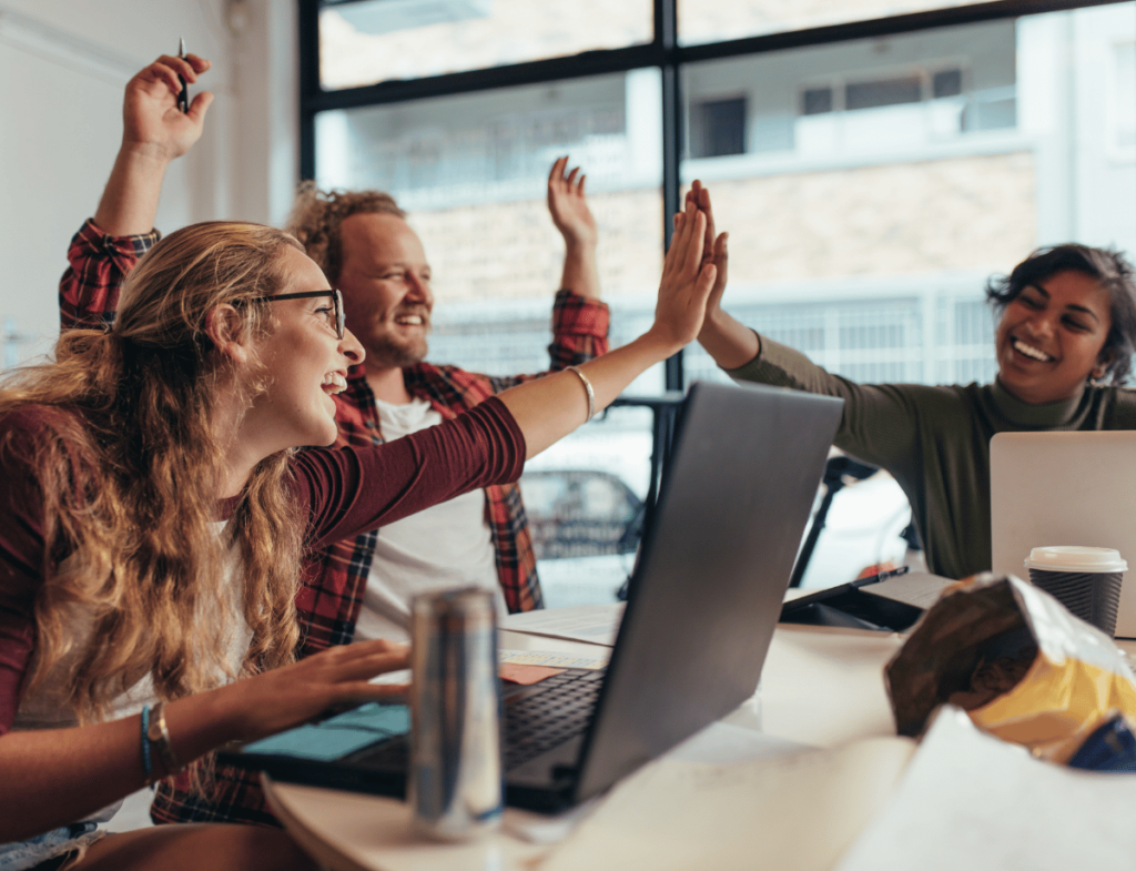 A group of 3 individuals sitting around a table looking at a laptop while 2 of the individuals engage in a high-five